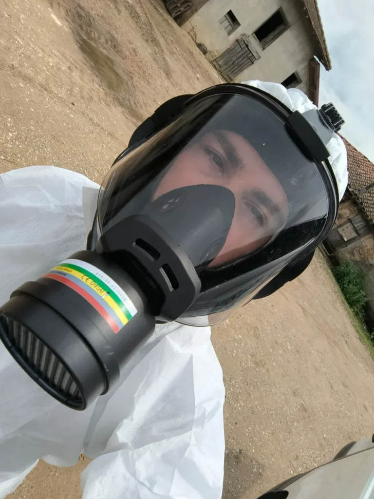 Personne portant une combinaison blanche et un masque à gaz intégral avec filtre, en extérieur, sur un terrain vague avec des bâtiments en arrière-plan.